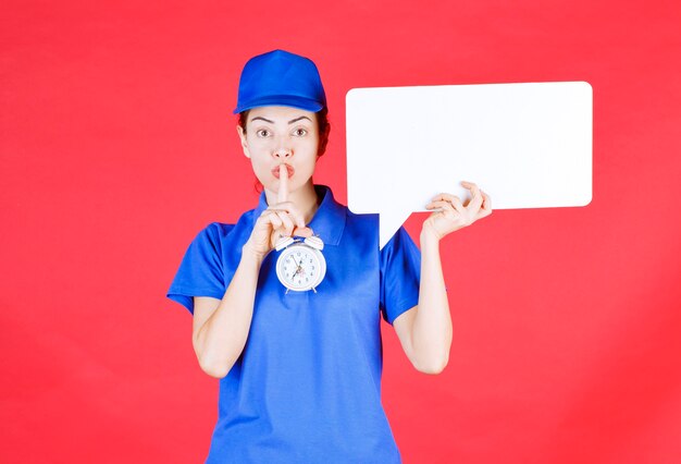 Guía femenina en uniforme azul sosteniendo un tablero de información rectangular blanco con un reloj despertador y pidiendo silencio.