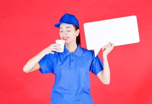 Guía femenina en uniforme azul sosteniendo un tablero de información rectangular blanco y tomando una taza de bebida desechable.