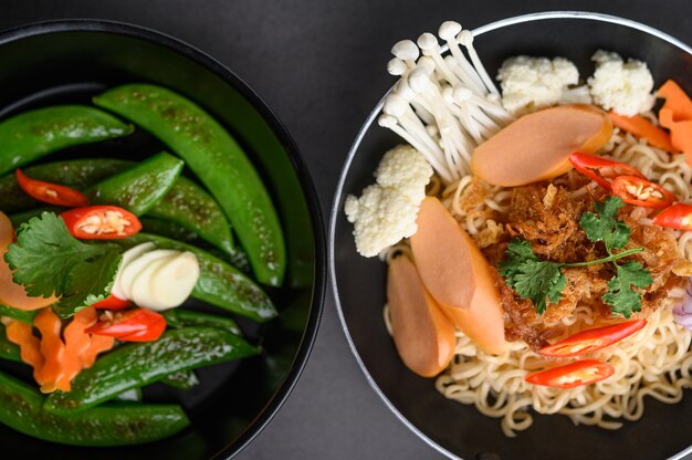 Guisantes salteados en una sartén y fideos picantes en el recipiente con ingredientes en la superficie de cemento negro.