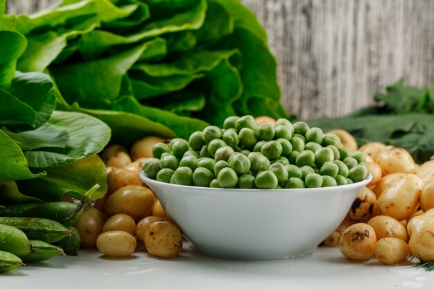 Guisantes con vainas verdes, espinacas, acedera, lechuga en un recipiente en la pared blanca y sucia, vista lateral.