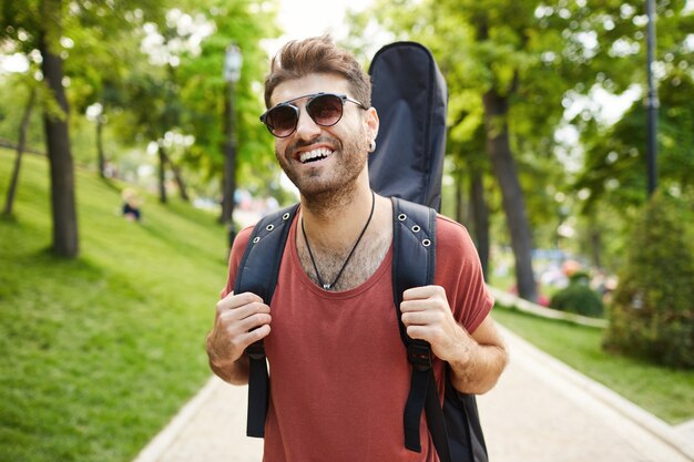 Guitarrista sonriente despreocupado, chico con guitarra caminando en el parque feliz