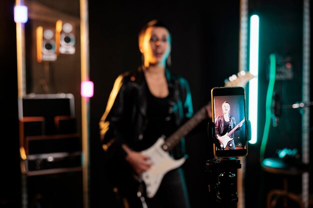 Guitarrista sosteniendo guitarra eléctrica tocando una canción de heavy metal mientras graba una actuación con el teléfono durante una sesión de música en un estudio de audio. Mujer músico trabajando en un nuevo álbum grunge antes de la gira