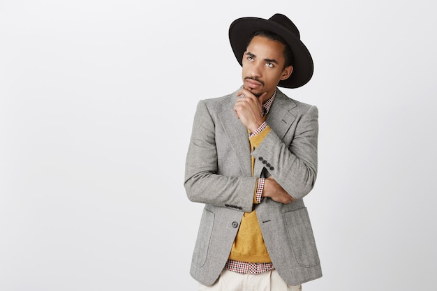 Guy se centró en pensamientos, tomando decisiones difíciles. Freelancer masculino serio concentrado de piel oscura con sombrero negro y elegante chaqueta, tocando la barbilla, mirando hacia arriba con expresión pensativa preocupada