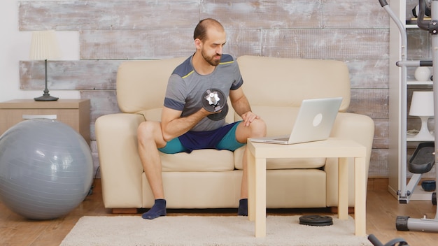 Guy entrenamiento de bíceps con mancuernas después del entrenamiento en línea en la computadora portátil.