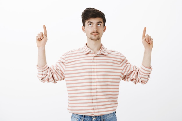 Guy parado, preocupado por la caída de rocas. Retrato de chico europeo nervioso intenso con bigote, levantando los dedos índices, mirando y apuntando hacia arriba, interesándose por lo que sucede hacia arriba