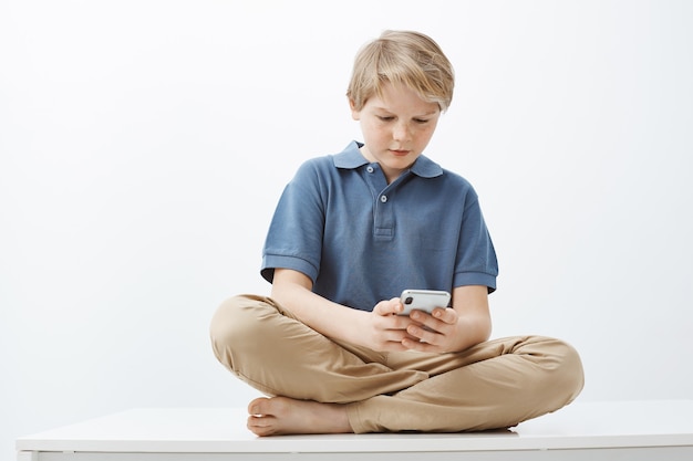 Guy se siente intenso jugando al juego favorito en el teléfono inteligente. Grave niño bonito con cabello rubio sentado en el piso con los pies cruzados, sosteniendo el teléfono y mirando la pantalla del dispositivo