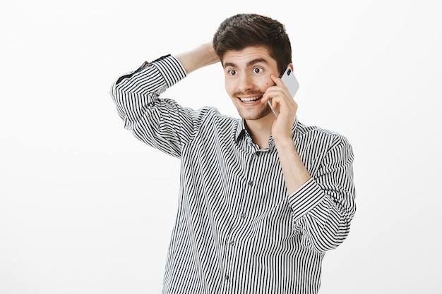 Guy sorprendido con la llamada. Retrato de alegre alegre joven barbudo europeo, rascándose la cabeza y hablando por teléfono inteligente, sonriendo, sintiéndose incómodo y tímido