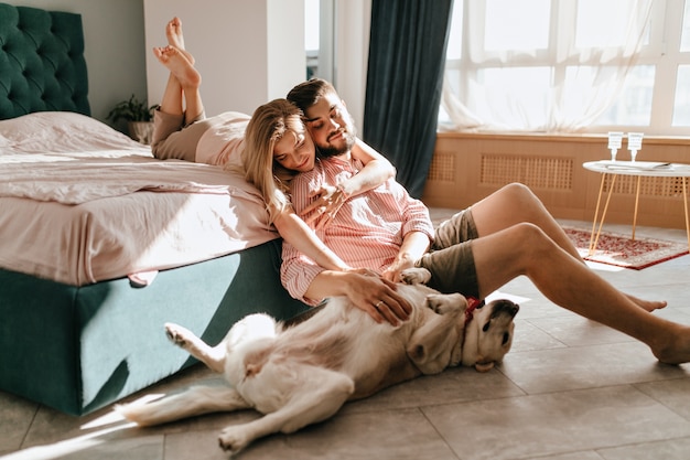 Guy y su novia descansan en el dormitorio. Feliz pareja mirando amorosamente a su mascota que quiere jugar.