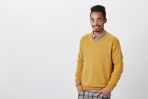 Guy sueña con convertirse en un médico famoso. Amistoso estudiante afroamericano ordinario de buen aspecto en jersey amarillo tomados de la mano en los bolsillos y sonriendo cortésmente, esperando el correo en la oficina de correos