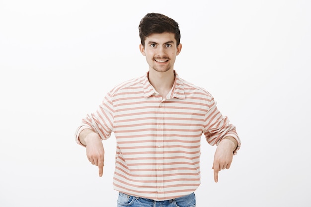 Guy sugiere la mejor oferta. Retrato de hombre barbudo intrigado complacido en camisa a rayas, apuntando hacia abajo