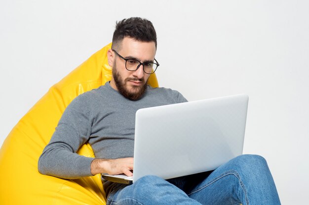 Guy trabaja con la computadora portátil mientras está sentado en una silla amarilla del puf