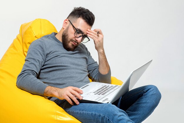 Guy trabaja con la computadora portátil mientras está sentado en una silla amarilla del puf