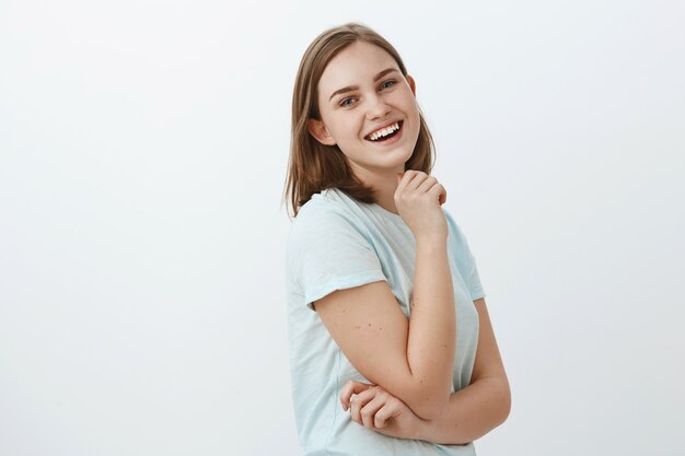 Hábil, creativa y ambiciosa mujer europea en camiseta de moda de pie en el perfil sobre la pared blanca girando con una sonrisa satisfecha, feliz y segura de sí misma, sosteniendo la mano en la barbilla