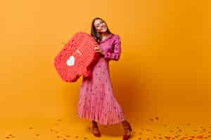 Foto gratuita en la habitación con pared de cartón naranja hay una elegante mujer de cabello castaño con falda larga, sosteniendo un gran cartel de instagram. foto tomada en colores naranja y rosa.