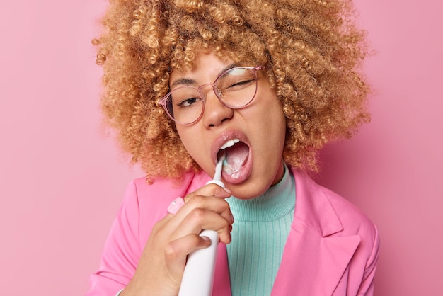 Hábitos y procedimientos de higiene diarios concepto Mujer hermosa joven rubia de pelo rizado limpia los dientes con cepillo de dientes eléctrico mantiene la boca abierta usa gafas y poses de chaqueta elegante interior