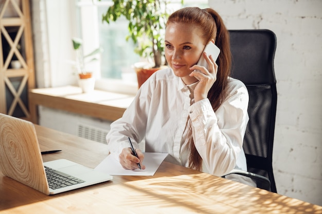 Hablando por telefono. Mujer joven caucásica en traje de negocios trabajando en oficina. Joven empresaria, gerente haciendo tareas con teléfono inteligente, computadora portátil, tableta tiene conferencia en línea. Concepto de finanzas, trabajo.