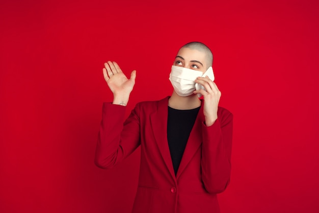 Hablando por telefono. Retrato de joven mujer calva caucásica aislada en la pared roja. Modelo de mujer hermosa en guantes, mascarilla. Las emociones humanas, la expresión facial, las ventas, el concepto publicitario.