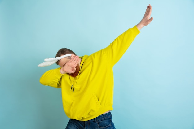 Haciendo dab. Hombre caucásico como un conejito de Pascua con ropa casual brillante sobre fondo azul de estudio. Felices saludos de pascua. Concepto de emociones humanas, expresión facial, vacaciones. Copyspace.