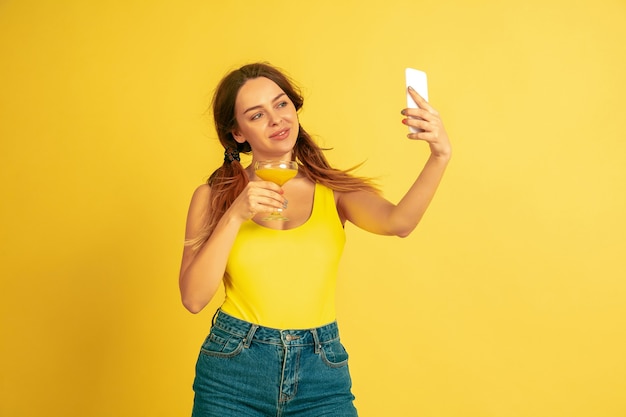 Haciendo selfie, vlog, sonriendo. Retrato de mujer caucásica sobre fondo amarillo de estudio. Modelo de mujer hermosa. Concepto de emociones humanas, expresión facial, ventas, publicidad. Verano, viajes, resort.