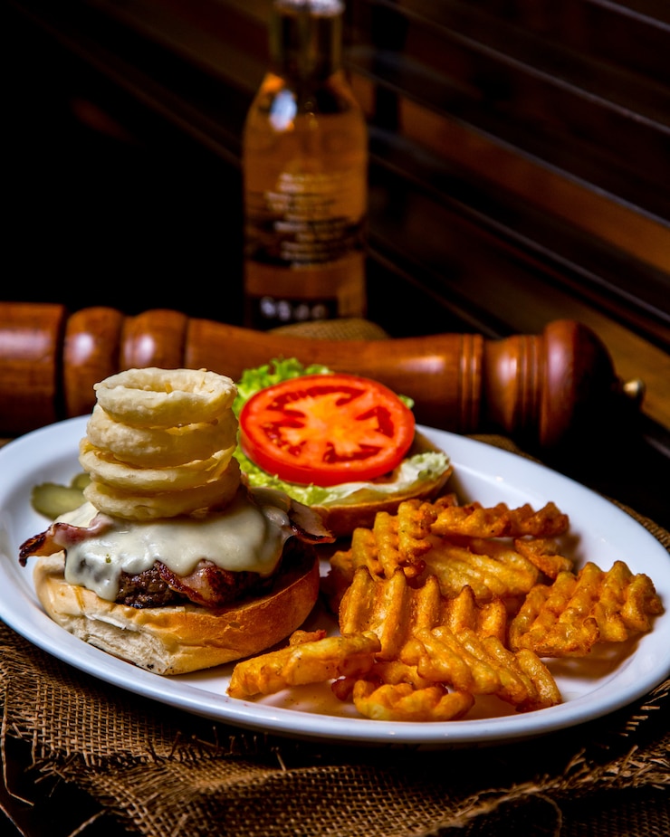 Foto gratuito hamburguesa abierta con carne, salsa de queso, aros de cebolla y tomate con papas fritas en un plato sobre una mesa de madera