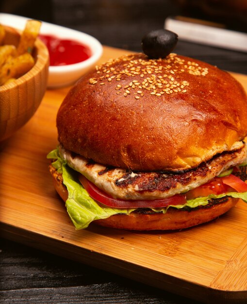 Hamburguesa de carne de pollo con rodajas de tomate y lechuga servida con papas fritas, salsa de tomate y mayonesa en una tabla de madera