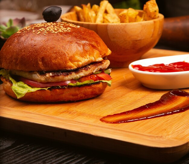 Hamburguesa de carne de pollo con tomate y lechuga en el interior y papas fritas servidas con aceituna negra y salsa de tomate en una bandeja de madera