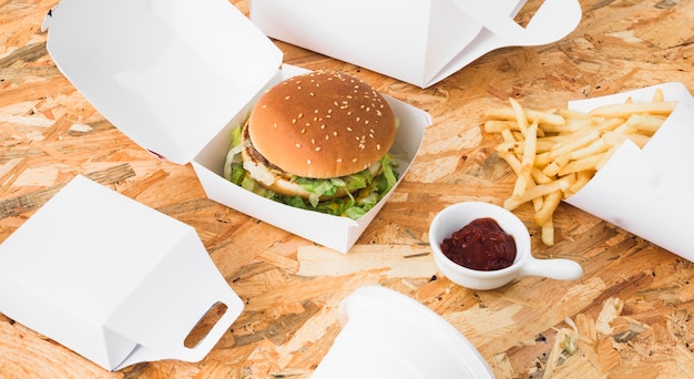 Foto gratuita hamburguesa; papas fritas y paquete de alimentos se burlan sobre fondo de madera