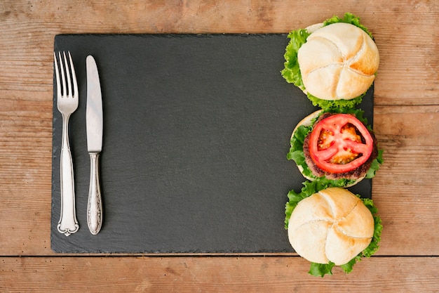 Foto gratuita hamburguesa plana en pizarra con cubiertos.