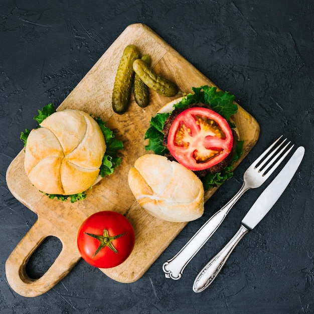 Hamburguesa plana sobre tabla de madera con cubiertos.