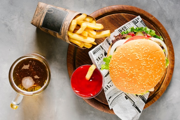 Hamburguesa plana sobre tabla de madera con papas fritas y cerveza.