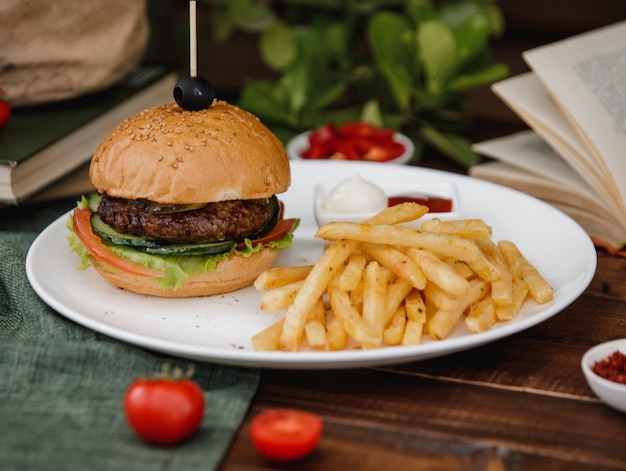 Una hamburguesa servida con papas fritas y salsas en un plato sobre una mesa rústica.