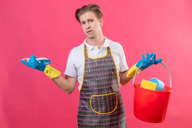 Hansdome joven hombre vestido con delantal y guantes de goma sosteniendo un balde con herramientas de limpieza y spray de limpieza