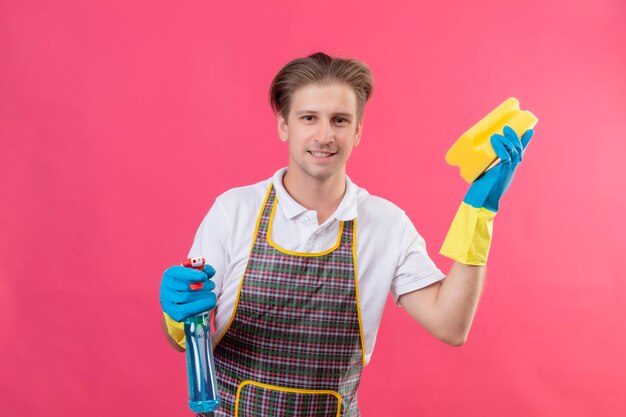 Hansdome joven hombre vestido con delantal y guantes de goma sosteniendo spray de limpieza y una esponja con sonrisa de confianza listo para cean de pie sobre la pared rosa 2