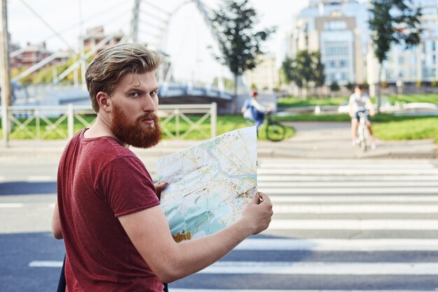 Hansome hombre con barba grande en camiseta roja camina en la ciudad para aprender más sobre ella