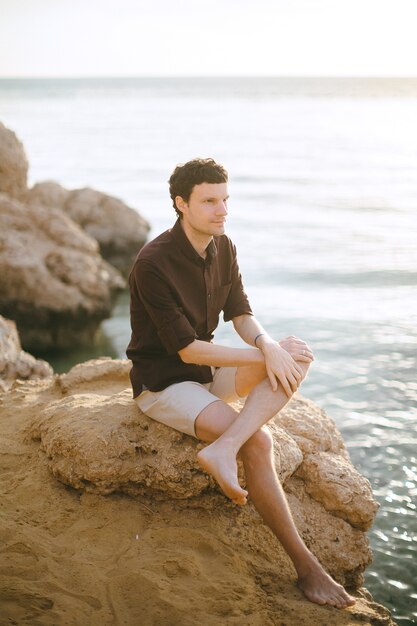 Hansome hombre de camisa negra sentado en las rocas y mirando lejos en la orilla del mar durante el día.