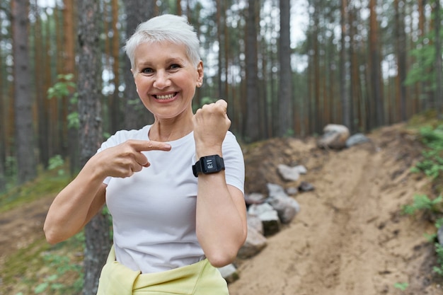 Happy fit mujer jubilada en ropa deportiva sonriendo ampliamente apuntando a la pantalla del reloj inteligente de muñeca