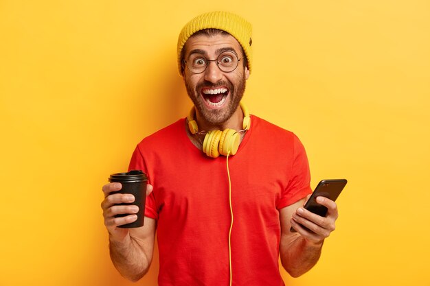 Happy hipster crea un nuevo perfil en las redes sociales, se ríe de la felicidad, sostiene un dispositivo electrónico moderno, bebe café