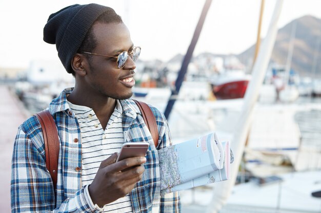 Happy te emocionó: mochilero afroamericano con elegantes gafas de sol y sombrero con un mapa de papel debajo del brazo, usando la aplicación de navegación en línea en el teléfono celular antes de su viaje