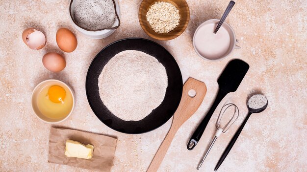 Harina en el plato; huevo; mantequilla; Leche; salvado de avena con espátula; Batidores y cuchara medidora sobre fondo texturizado.