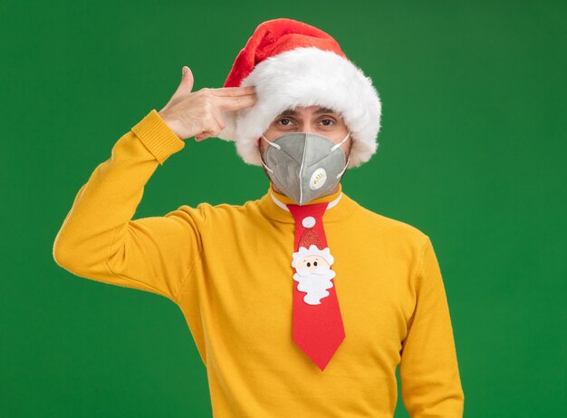 Harto joven caucásico con sombrero de navidad y corbata con máscara protectora haciendo gesto de suicidio aislado en la pared verde