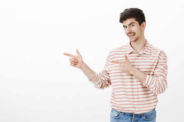 ¿Has conocido a mi amigo? Feliz y amigable compañero de trabajo con barba, señalando con gestos de pistola hacia la izquierda, pidiendo a sus amigos que visiten el bar, se relajen y se relajen, sonriendo con curiosidad sobre la pared gris