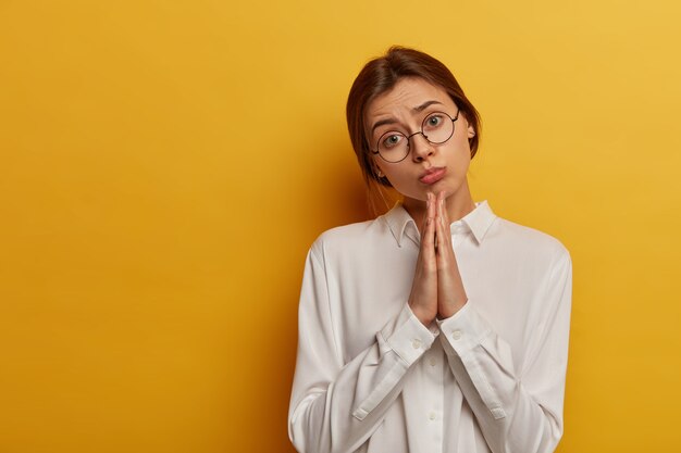 ¡Hazme un favor una vez más! Mujer joven y atractiva tiene cara triste, sostiene ambas palmas en gesto de oración, pide ayuda a un amigo, viste camisa blanca