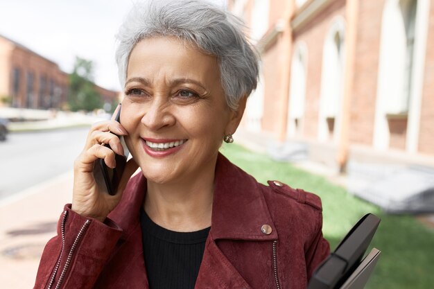 Headportrait de mujer de mediana edad con chaqueta posando en las calles de la ciudad con el móvil en la oreja