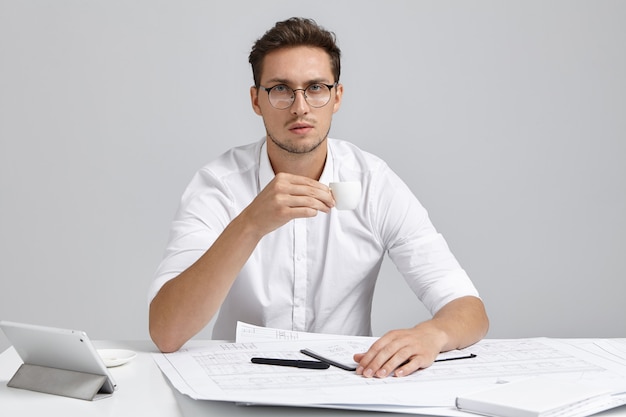 Headshotof atractivo joven constructor europeo cansado con barba sosteniendo una taza blanca, bebiendo espresso mientras trabaja toda la noche en orden urgente