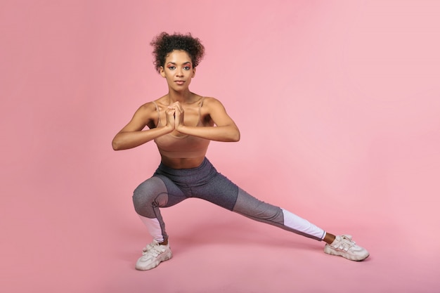 Hembra afroamericana confiada que hace ejercicios en estudio sobre fondo del pivk. Elegante atuendo de fitness.