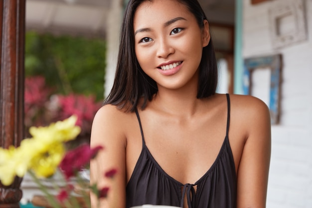 La hembra asiática alegre con piel sana descansa bien en un restaurante junto con un amigo, tiene una expresión soñadora, nota algo agradable por la ventana.