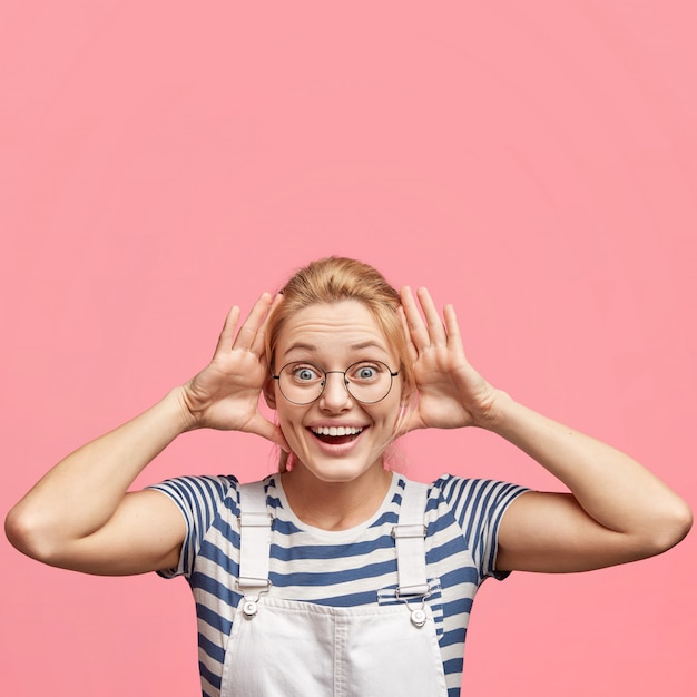 Hembra divertida llena de alegría con expresión alegre hace muecas, mantiene las manos cerca de las orejas, tiene una mirada feliz y aturdida, aislada sobre rosa