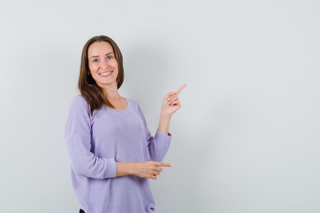 Foto gratuita hembra joven apuntando a un lado en blusa lila y mirando positivo