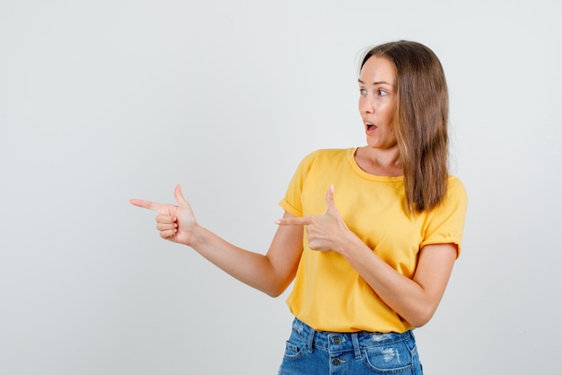 Hembra joven apuntando hacia el lado con gesto de pistola en camiseta, pantalones cortos y mirando alegre