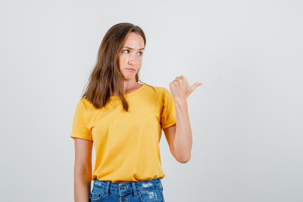 Hembra joven apuntando con el pulgar a un lado en camiseta, pantalones cortos y mirando decepcionado. vista frontal.